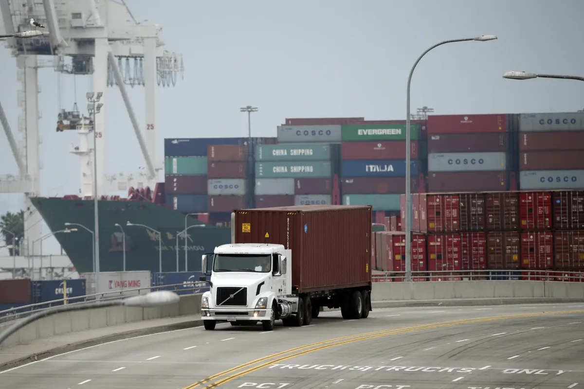 White tractor hauling an ocean container past marine-terminal cranes and container stacks